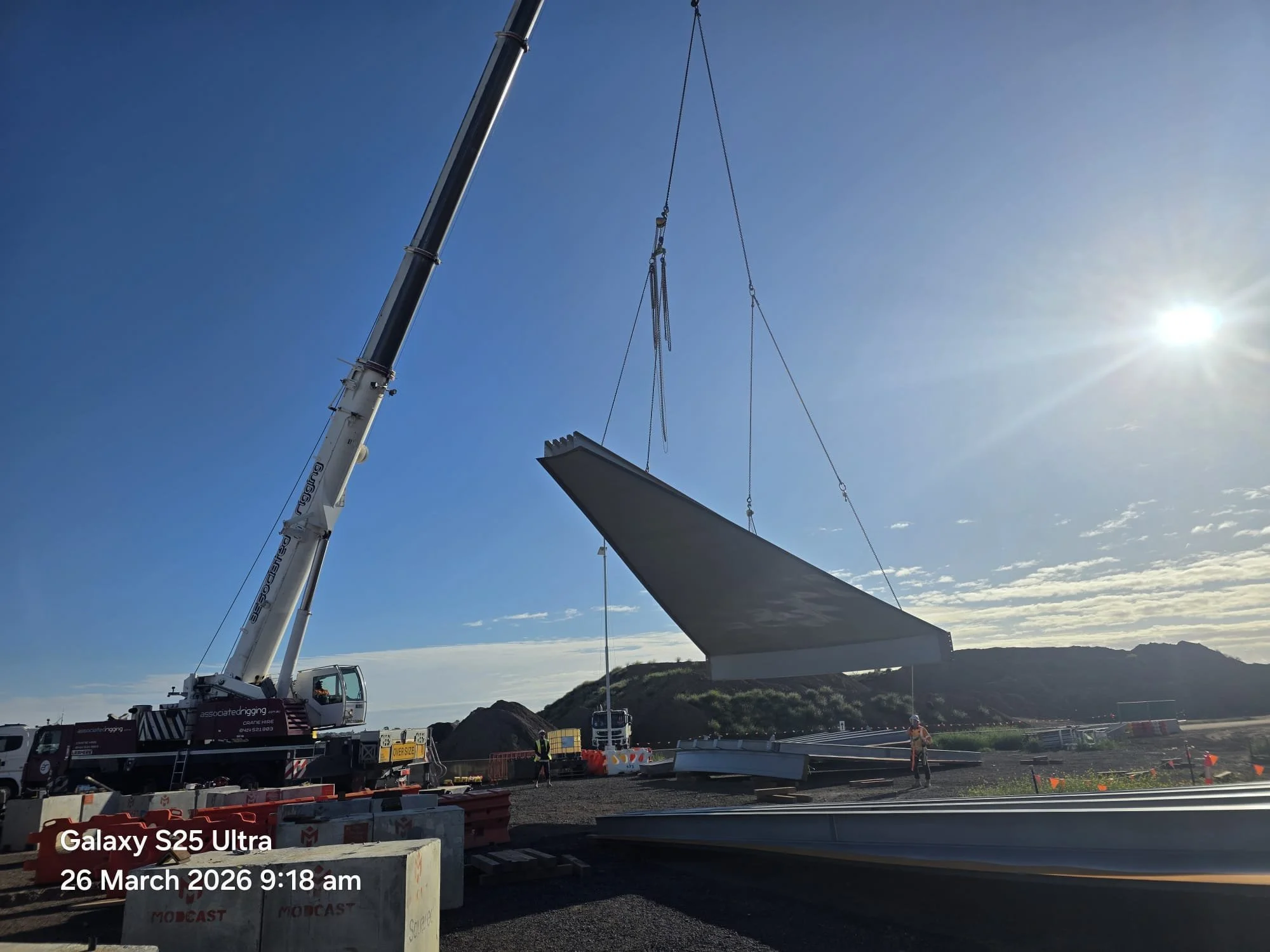 A construction crane lifting a large panel or piece of material at a construction site on a sunny day. Several construction workers and orange safety barriers are visible on the ground.