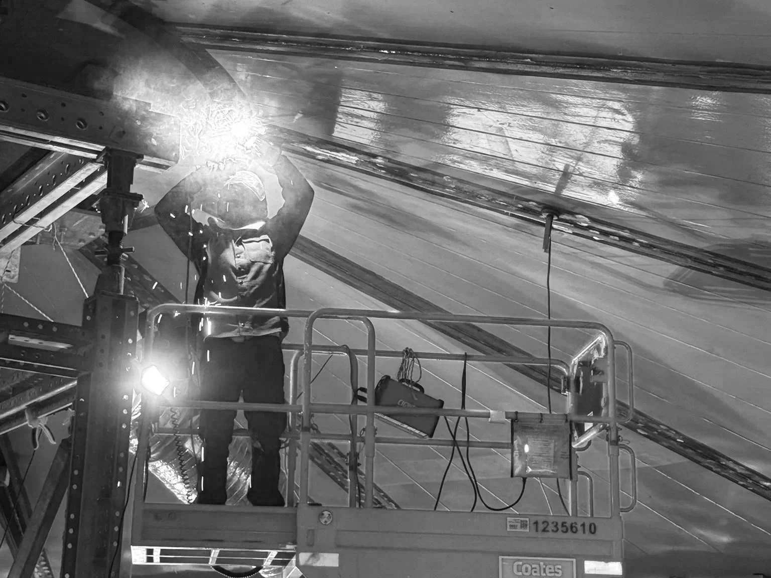 A worker in safety gear welding on a metal structure while standing on a platform in an industrial setting, with sparks and bright light from the welding process.
