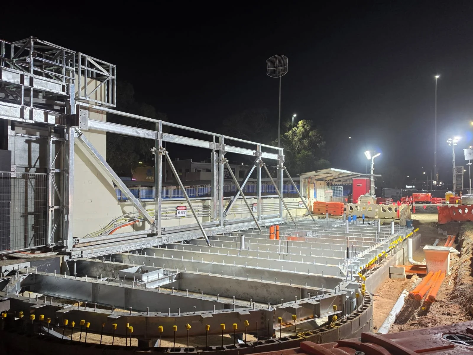 Nighttime construction site with metal framework, orange barriers, and bright floodlights illuminating the area.