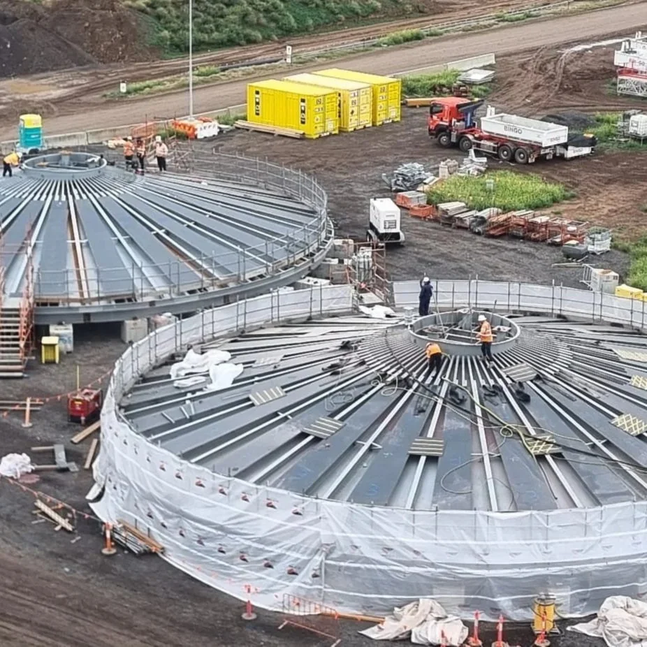 Construction workers assemble large, circular steel structures on a muddy construction site with equipment, portable toilets, and yellow storage containers in the background.