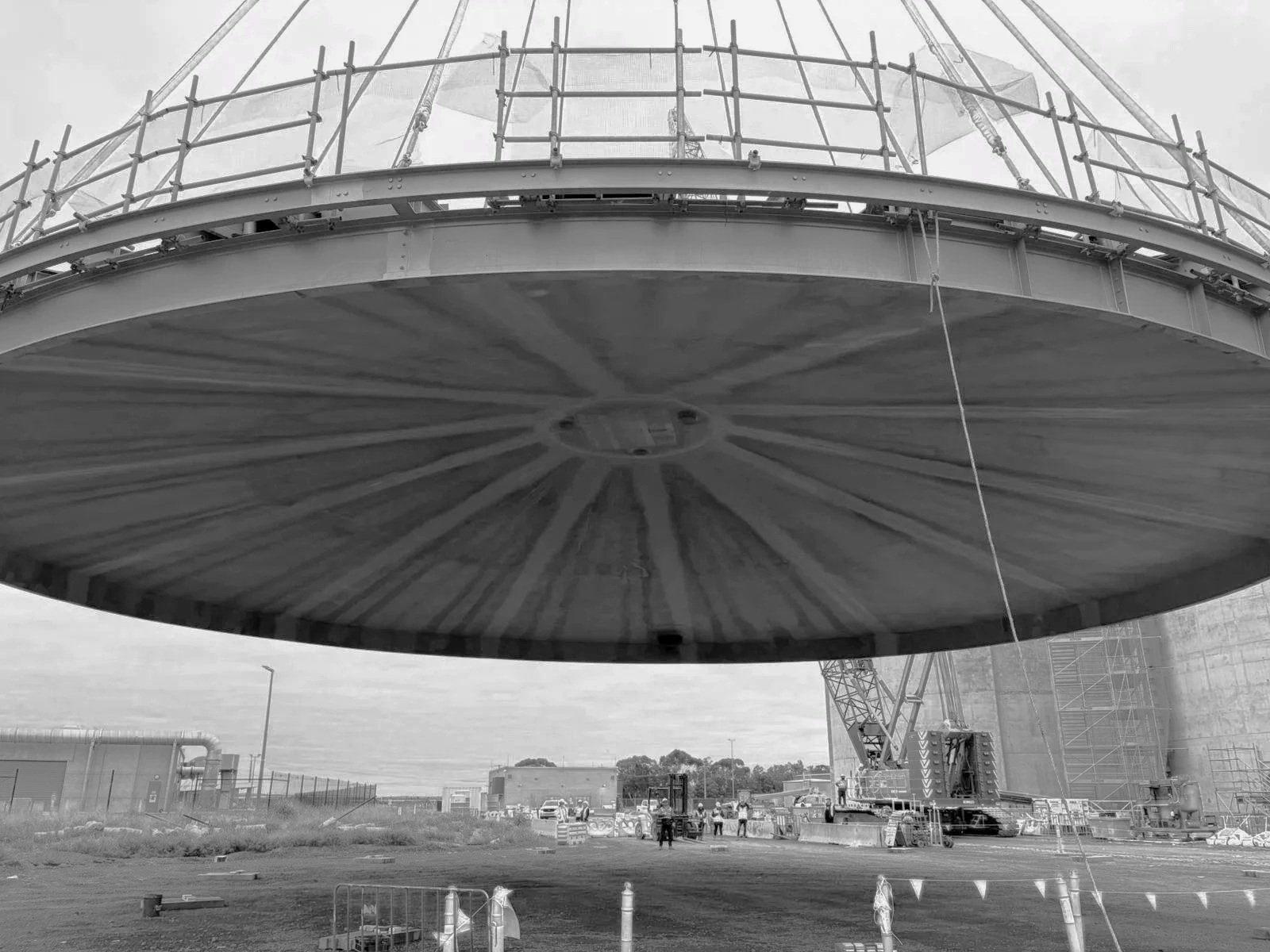 Large construction crane installing a giant domed structure, with workers on the ground nearby.