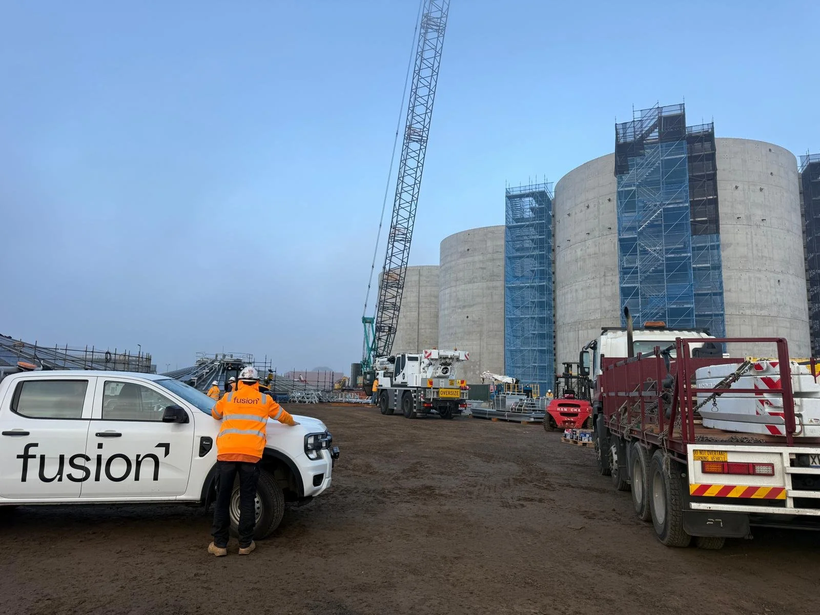 Construction site with large concrete silos, cranes, and construction workers in safety gear. Vehicles and scaffolding are present on the dirt ground.
