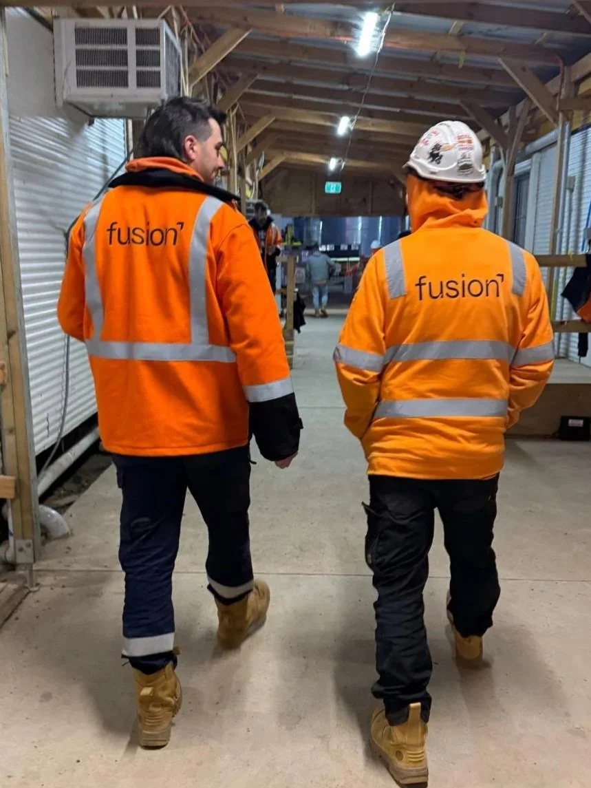 Two workers in orange high-visibility jackets with the company's name 'fusion' on the back are walking through an indoor construction site. They are wearing safety boots and one has a safety helmet. The space has wooden beams and metal wall panels.
