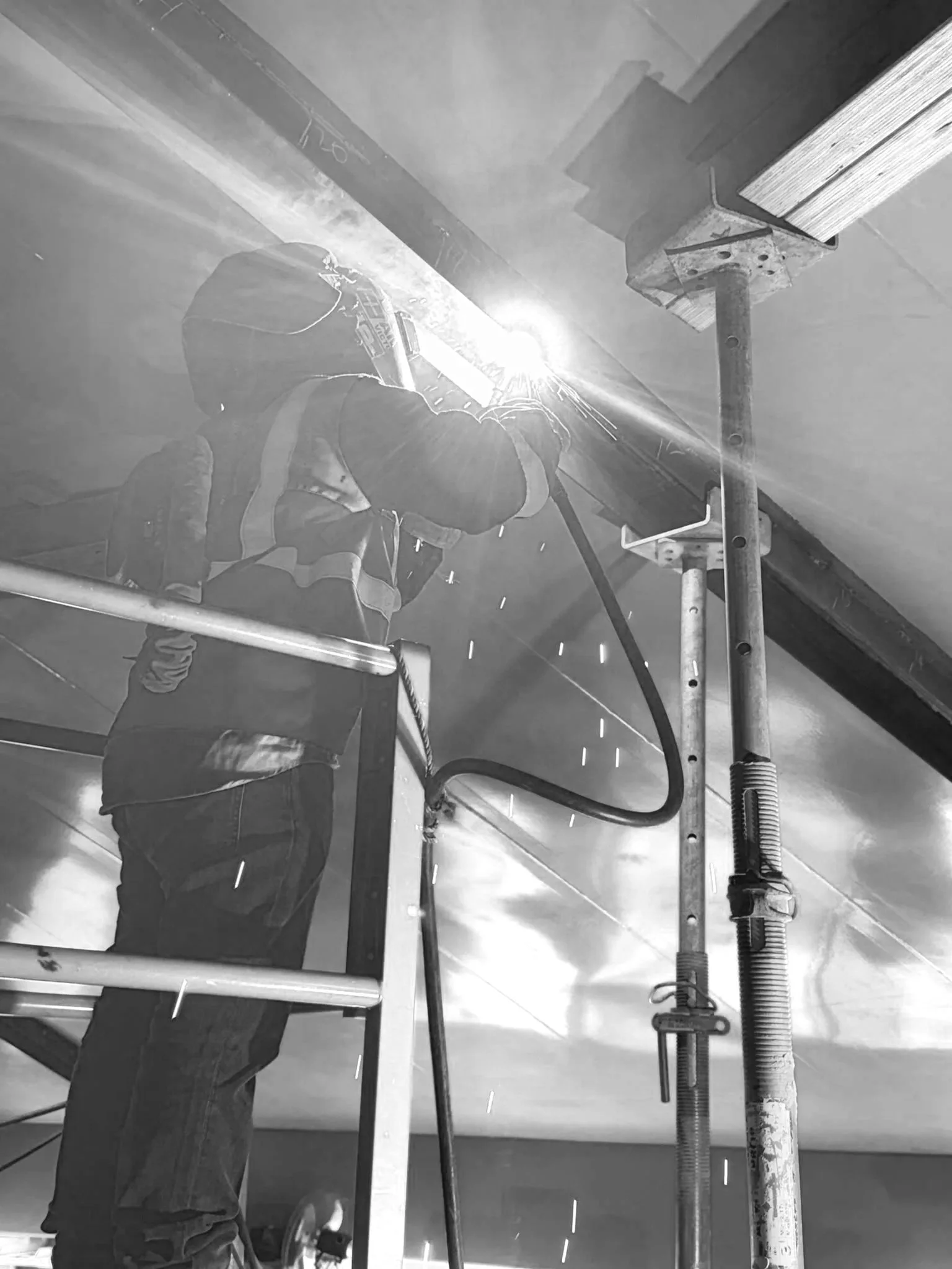 A worker welding metal on a construction site, with protective gear and bright sparks visible, using scaffolding and safety equipment.
