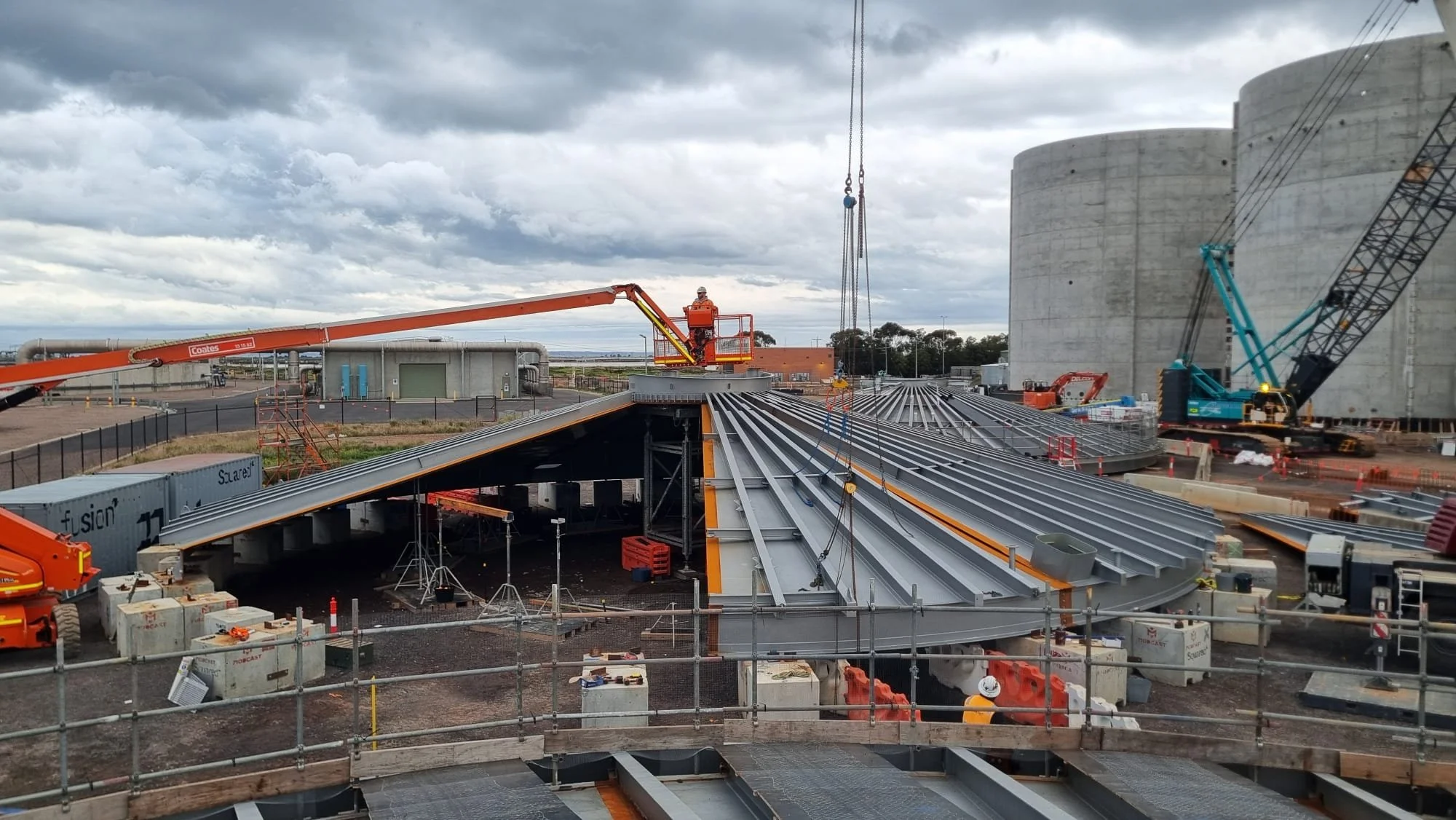 Construction site with large gray industrial storage tanks, cranes, and workers installing a metal roof structure on a building, under cloudy sky.