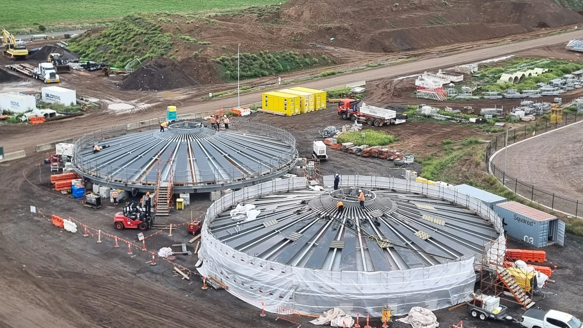 Construction site with large circular steel structures being assembled, workers in safety gear, construction equipment, and supplies, with dirt roads and grassy hills in the background.