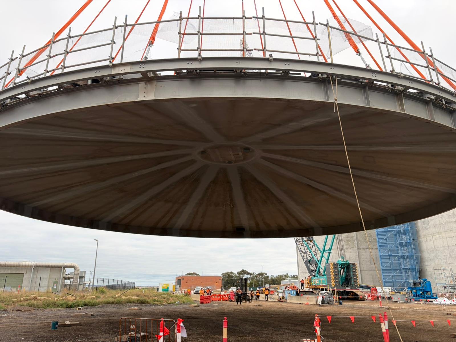 A large construction site with a massive, cylindrical structure being supported and assembled, with construction workers, cranes, and safety barriers visible.