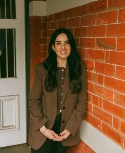 A woman with long dark hair, wearing a brown blazer, standing outdoors against a red brick wall.