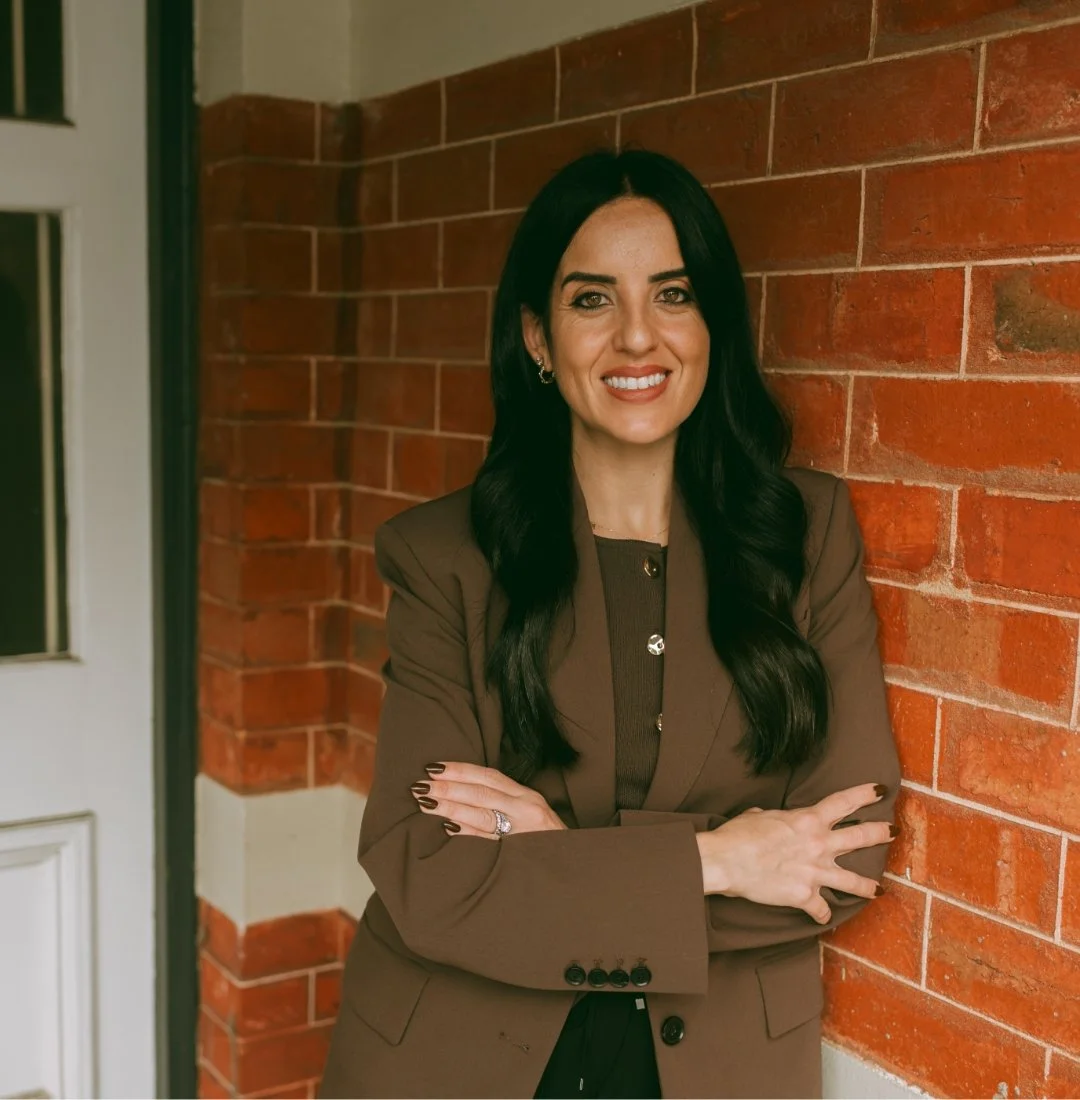 A woman with long black hair and light skin, wearing a brown blazer, standing with arms crossed in front of a brick wall, smiling.