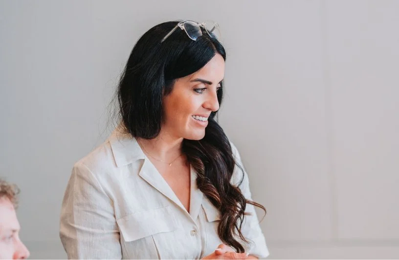 A smiling woman with long dark hair wearing glasses on her head and a light-colored shirt, looking to her right.