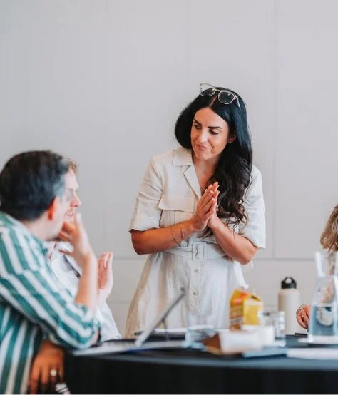 A woman standing and speaking to a group of people seated at a table in a meeting room.