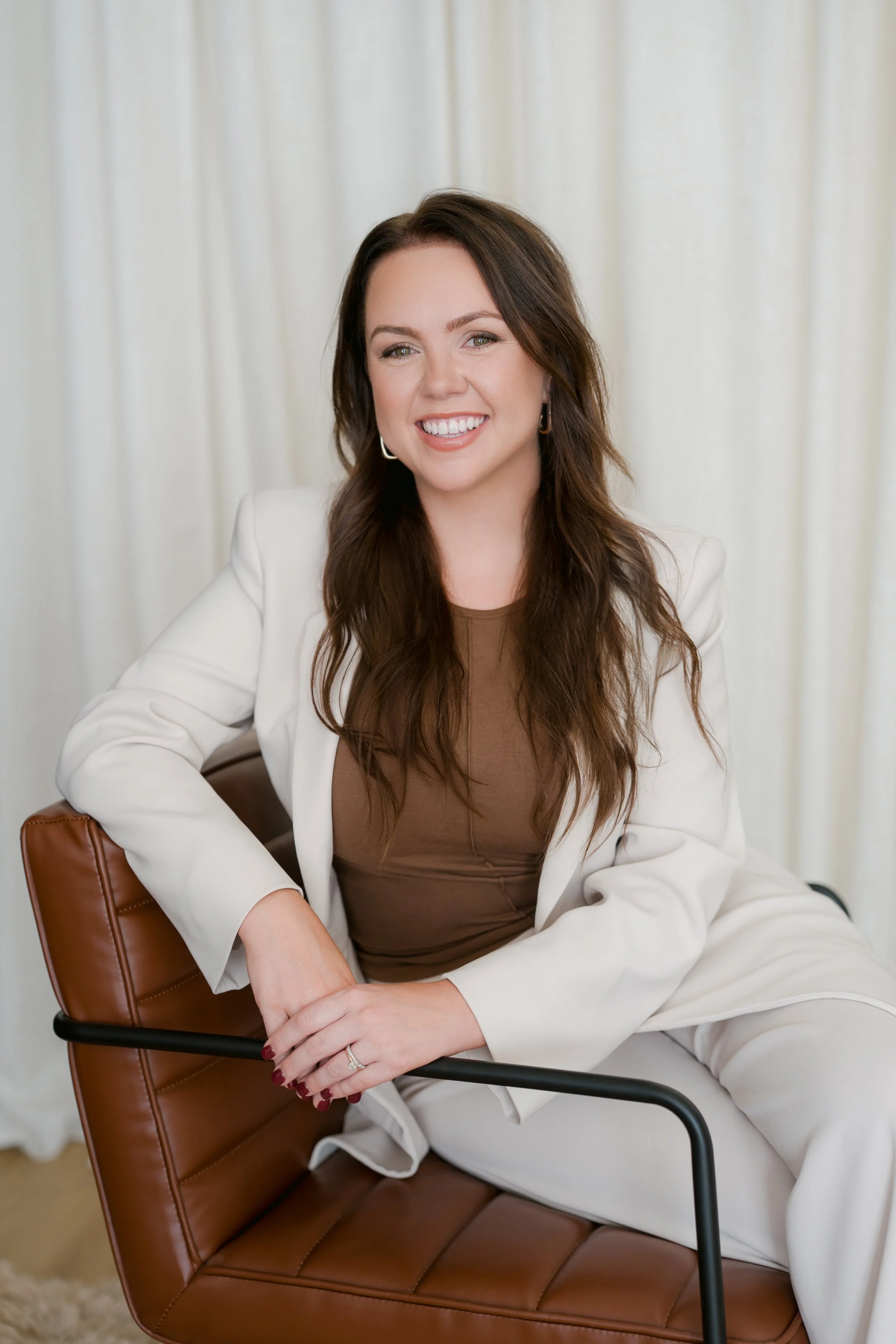 A young woman with brown hair and a white blazer smiling outdoors with blurred greenery and water in the background.