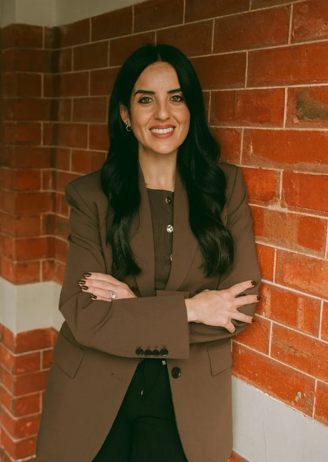 A woman with long dark hair, smiling, wearing a brown blazer and black pants, standing against a red brick wall with her arms crossed.