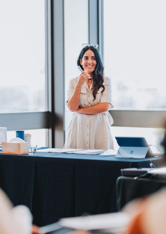 A woman with long dark hair, wearing glasses on her head and a beige dress, stands happily by a table in front of large windows, speaking on a phone in a bright, modern conference room.