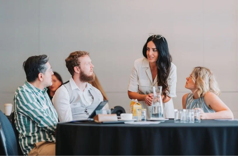 A woman standing and speaking to a group of three people sitting at a table during a business meeting or conference.