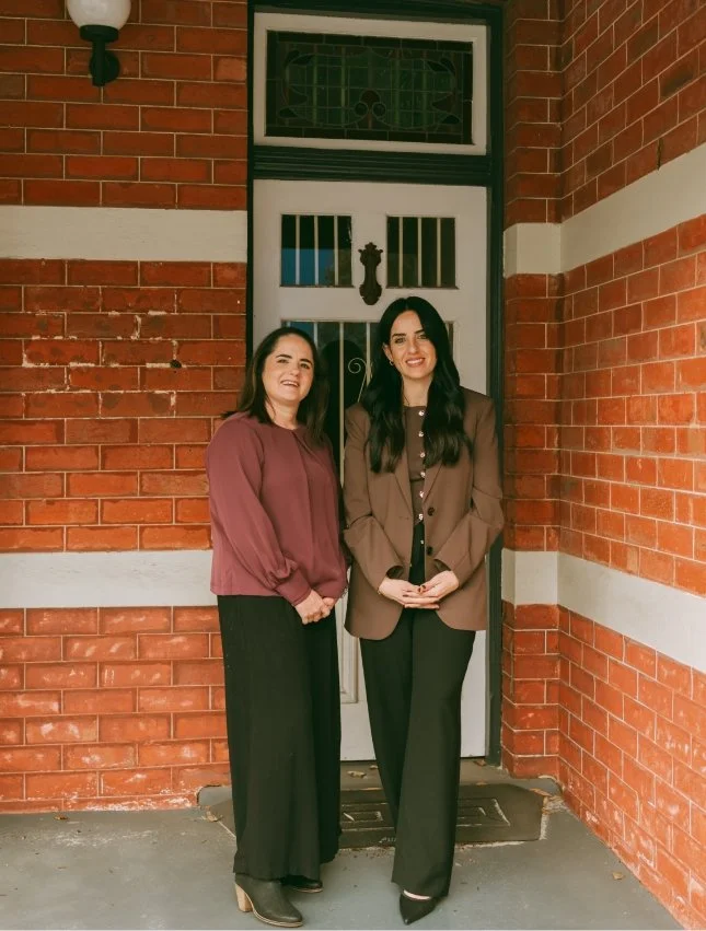 Two women are standing in front of a brick house with a white door and stained glass window. They are smiling and dressed in professional attire.
