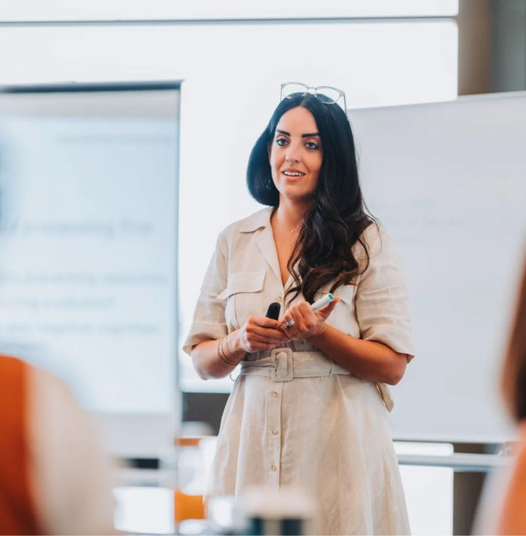 Woman with long dark hair, glasses on her head, wearing a beige dress, giving a presentation in a bright room with a whiteboard.