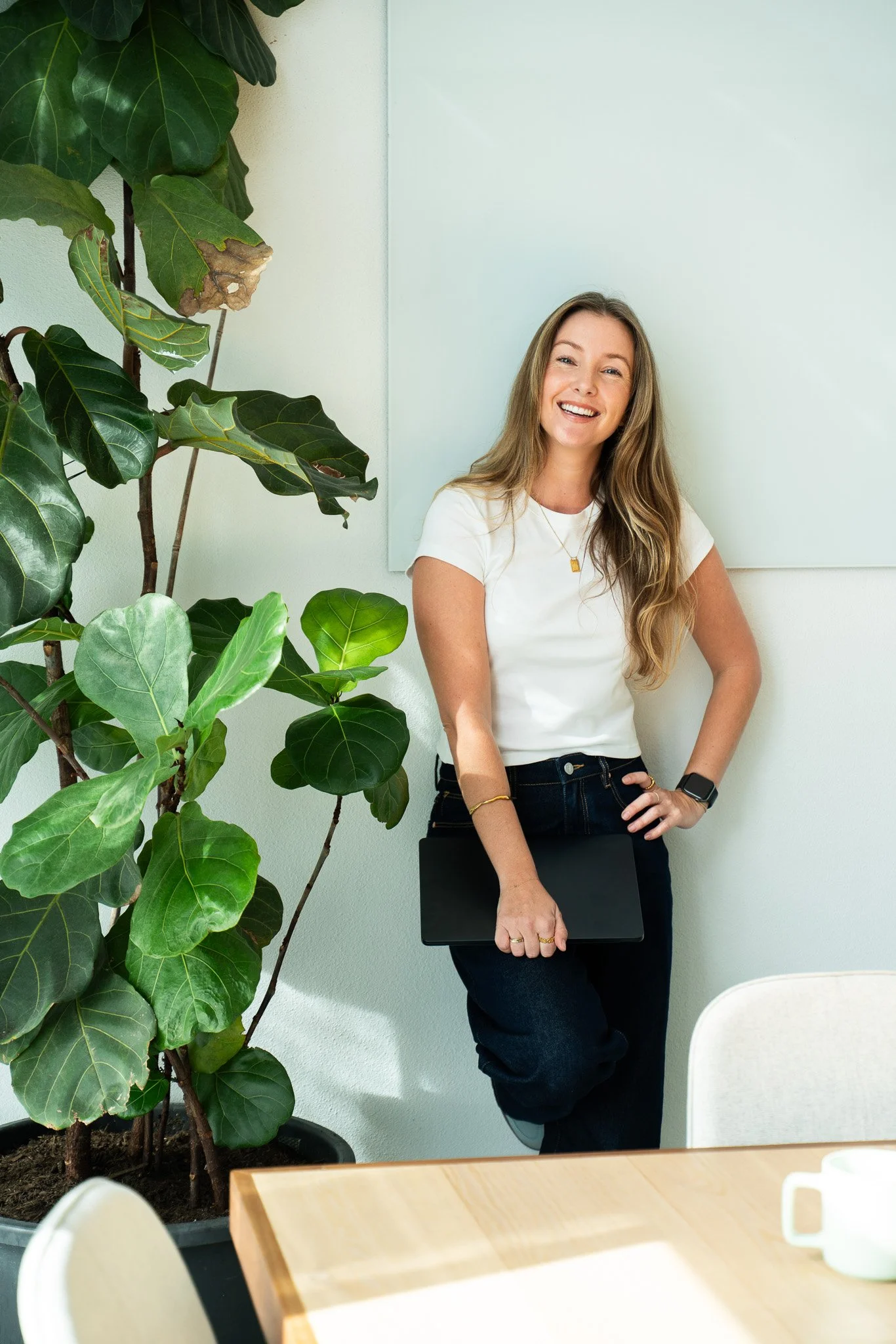 A woman with long wavy hair smiling, holding a closed laptop. She is wearing a white t-shirt, dark jeans, and a smartwatch. There is a large potted plant with big green leaves to her left and a light-colored wall with a whiteboard behind her. Part of a wooden table and a white chair are visible in the foreground.
