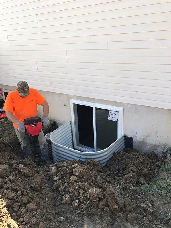 A worker in an orange shirt and camouflage hat installing a window well around a basement window outside a house. The area is dug out with dirt mounded around the edge.