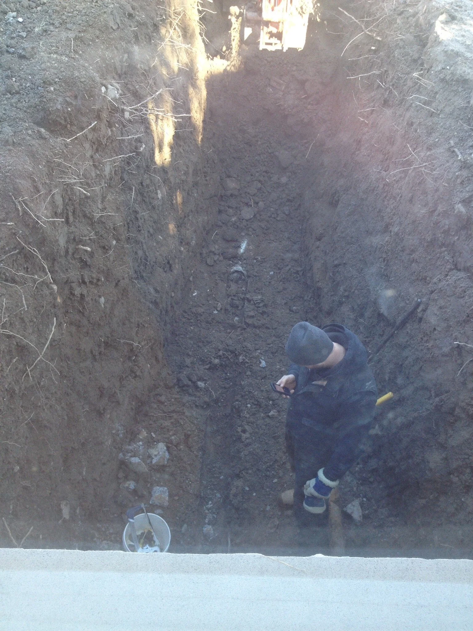 Person crouched in a deep hole, working on the earth, with a bucket and tools nearby, outdoors in a construction or excavation site.
