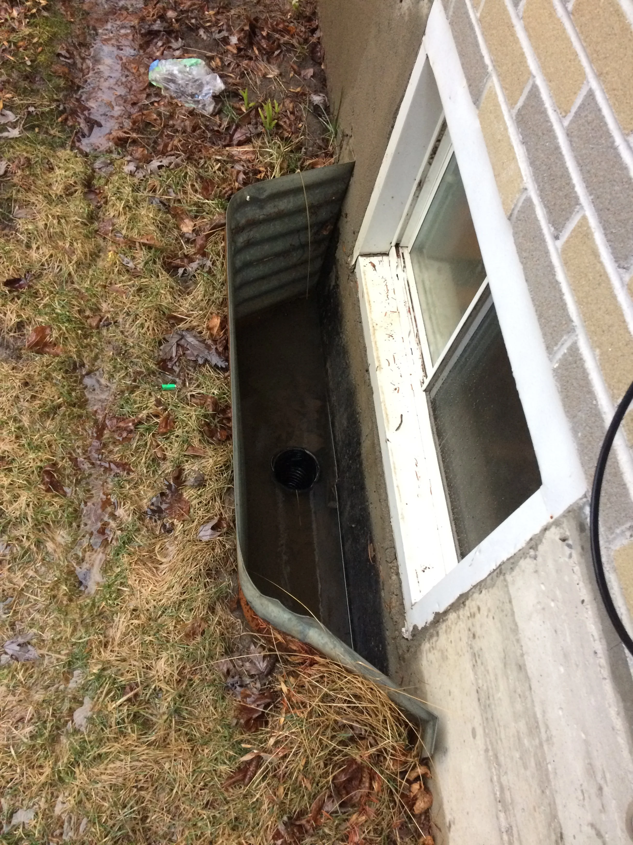 A partially disassembled metal window well cover lying on the ground outside a house, next to mulch and a brick wall.