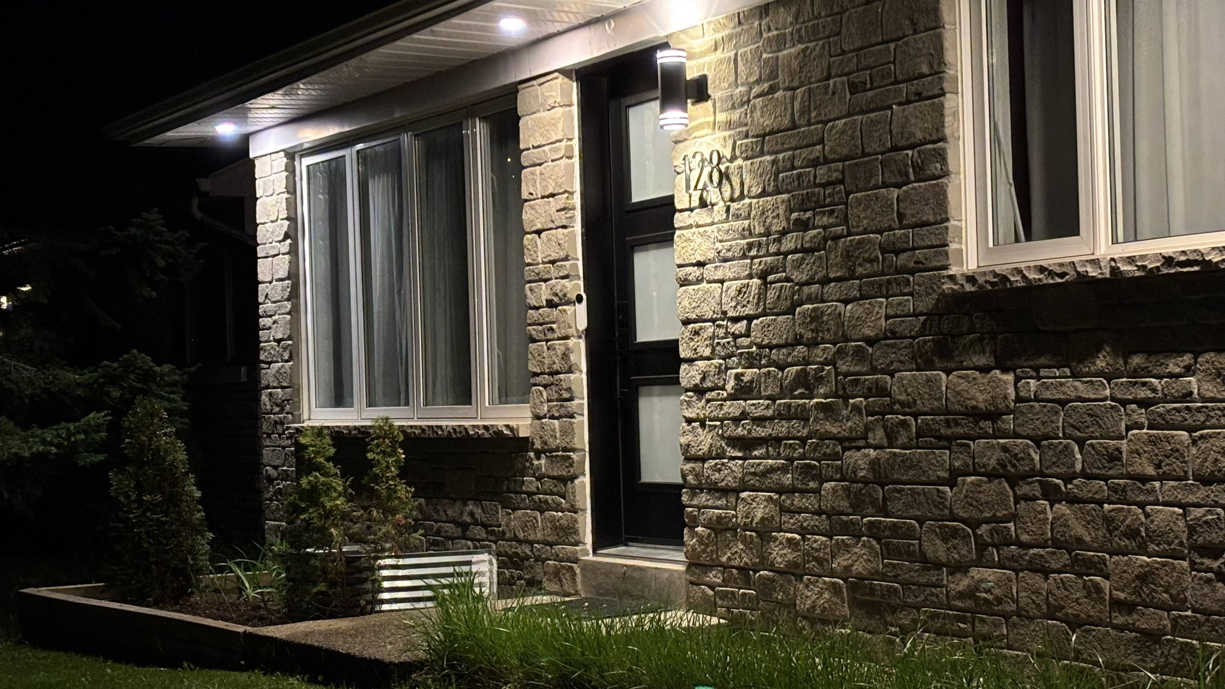 Nighttime view of a house with a brick exterior, illuminated porch light, and large glass windows.