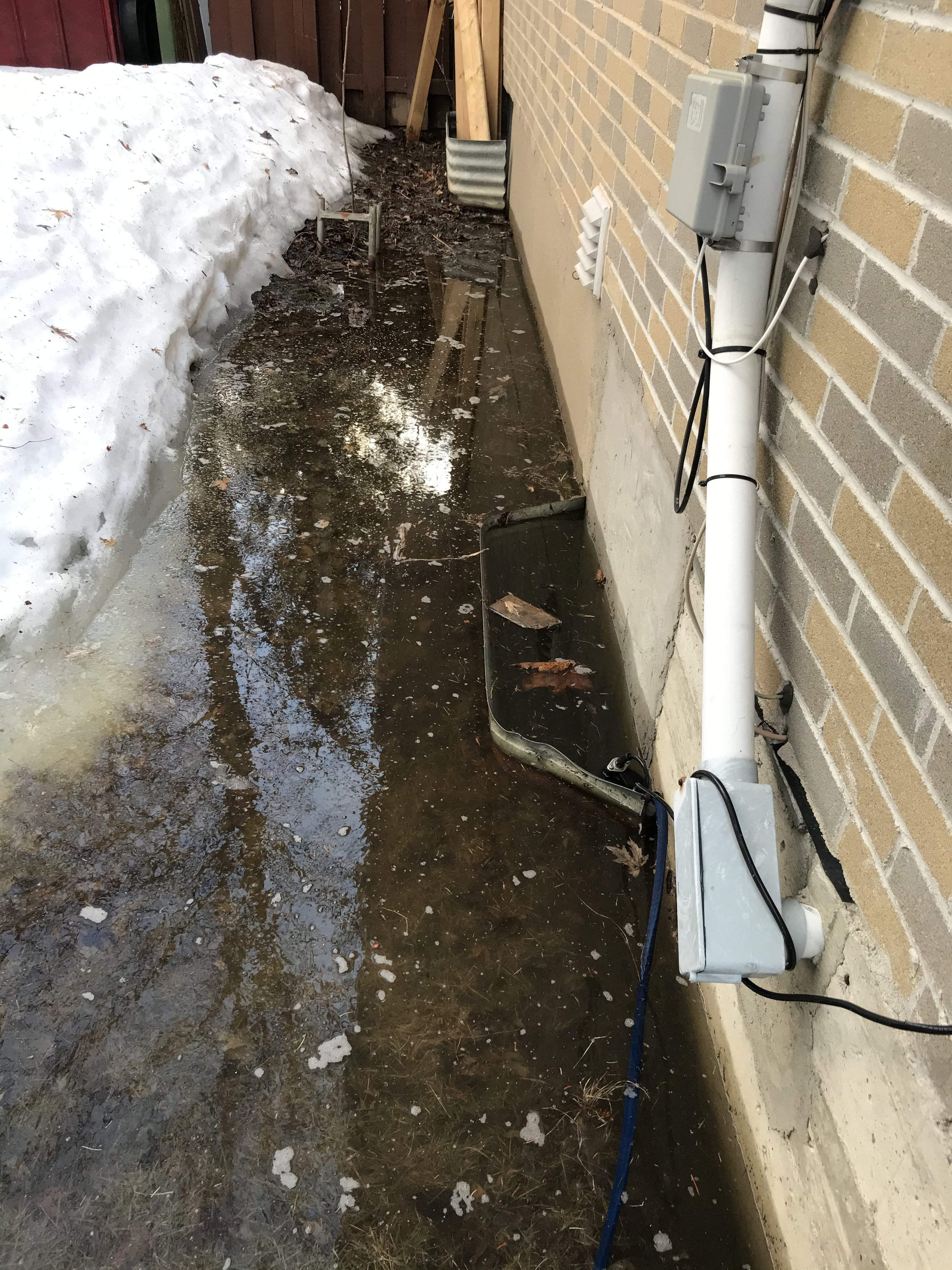 A small construction site with a partially completed concrete foundation, electrical equipment, and a pile of snow along the side of a brick house.