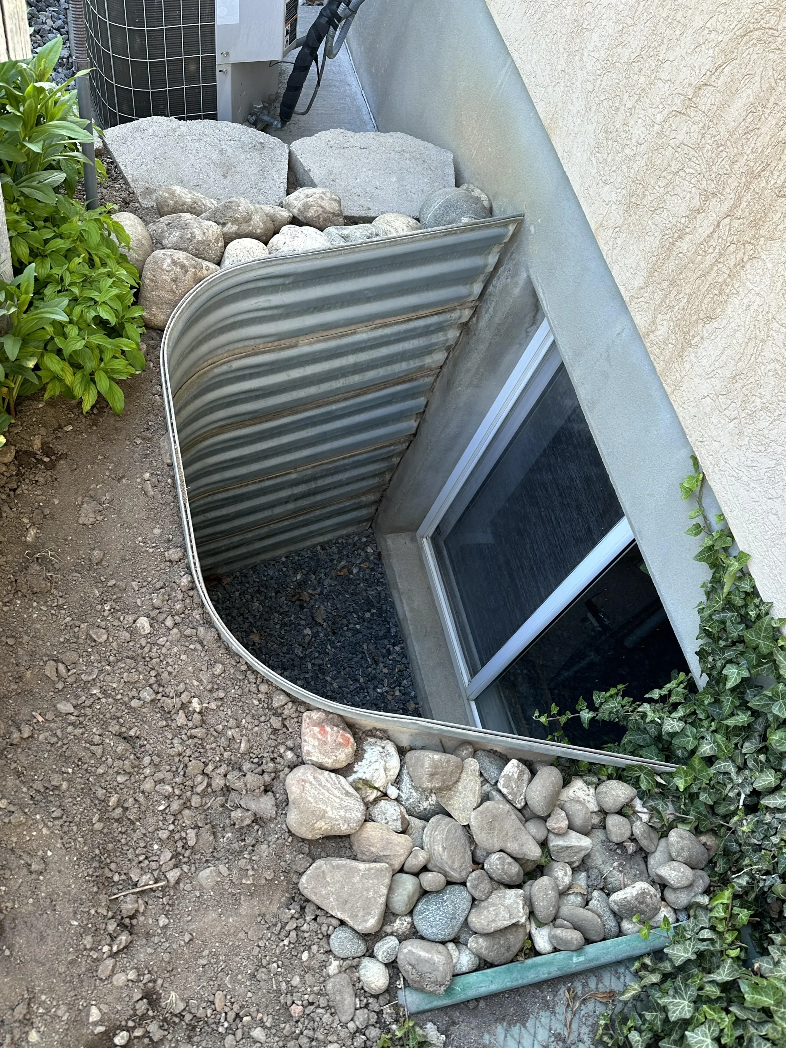 A small garden area next to a house foundation with a patch of dirt, a row of large and small rocks, a green plant on the left, and ivy on the right. There is a corrugated metal barrier lining the rocks, and part of an air conditioning unit is visible in the background.
