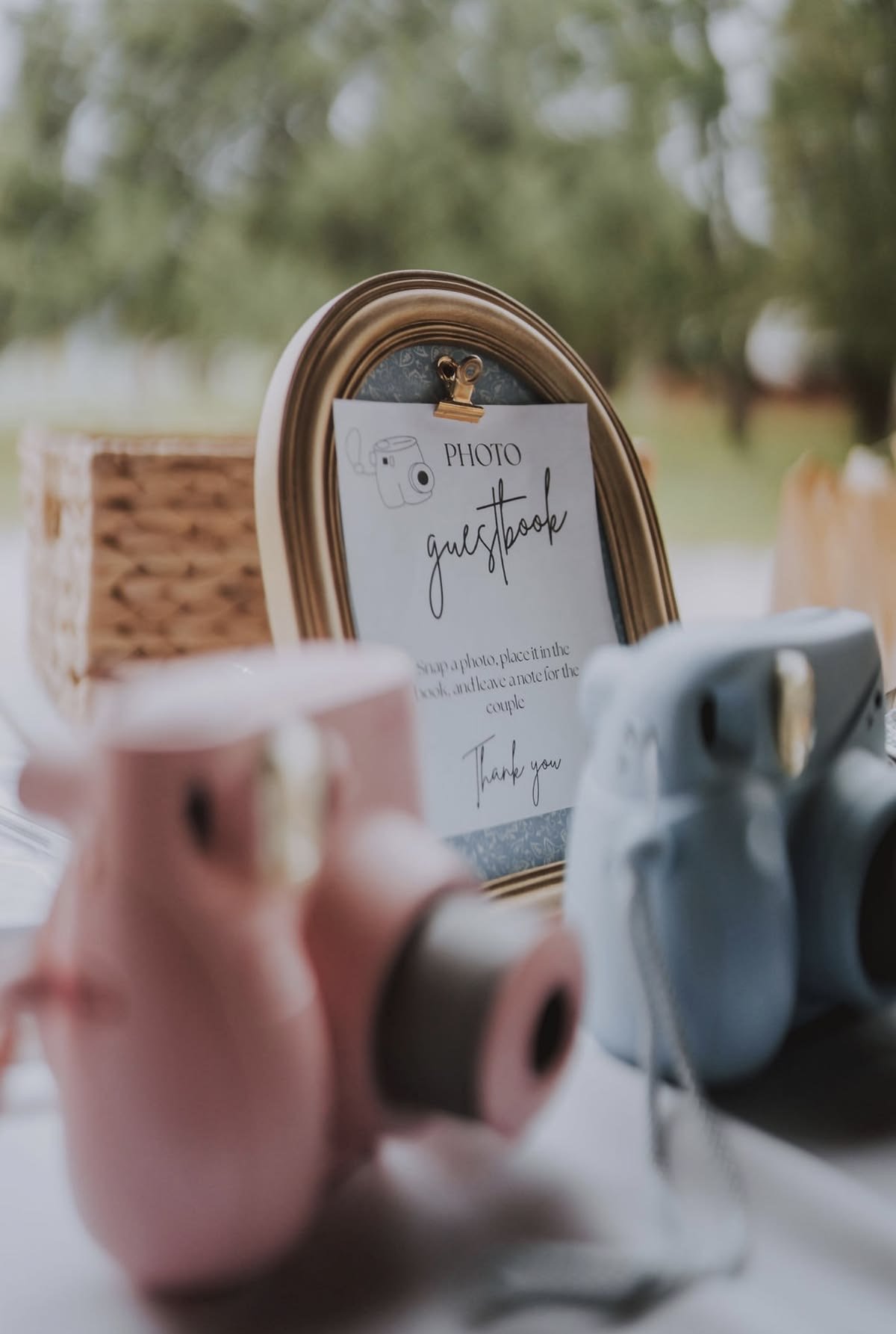 A photo of a guest book sign-in table at an outdoor event, with a framed sign that reads "Guestbook" and instructions to add a photo and note for a couple, surrounded by pink and blue ceramic teapots.
