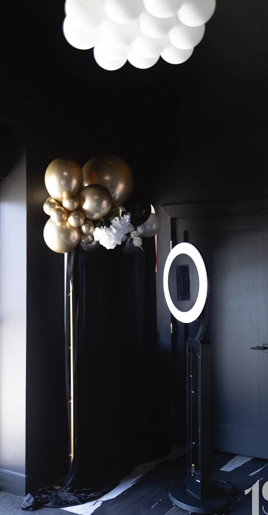Black room corner decorated with black, gold, and white balloons and flowers, and a photo booth with a ring light.