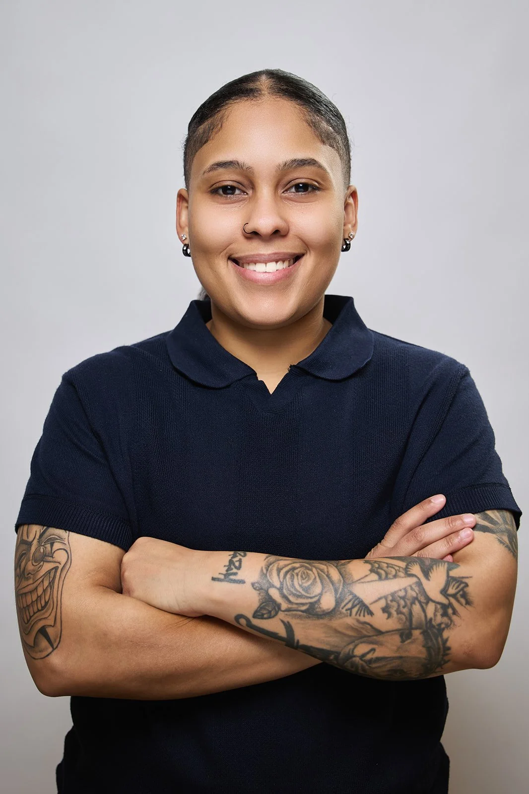 A young woman with short dark hair, tattoos on her arms, wearing a navy blue polo shirt, smiling with arms crossed against a plain grey background.