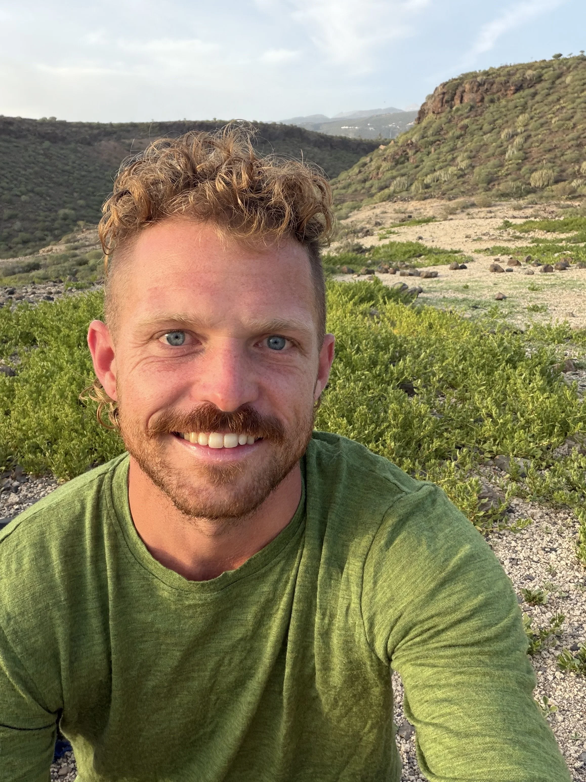 A portrait photo of Jungle Jovian smiling with curly blonde hair and a beard wearing a green shirt, outdoors with green hills and rocky terrain in the background.