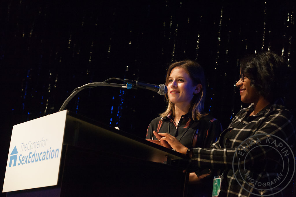 Two women speaking at a podium with a microphone during a conference about sex education, with a dark, glittery background.