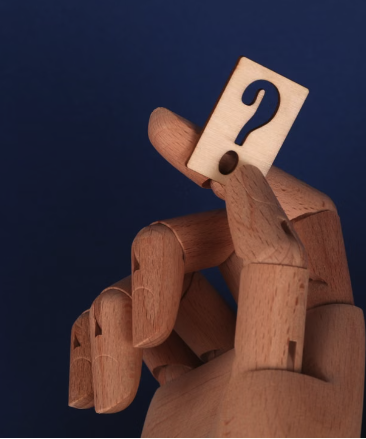 A wooden mannequin hand holding a small wooden block with a question mark on it, against a dark blue background.
