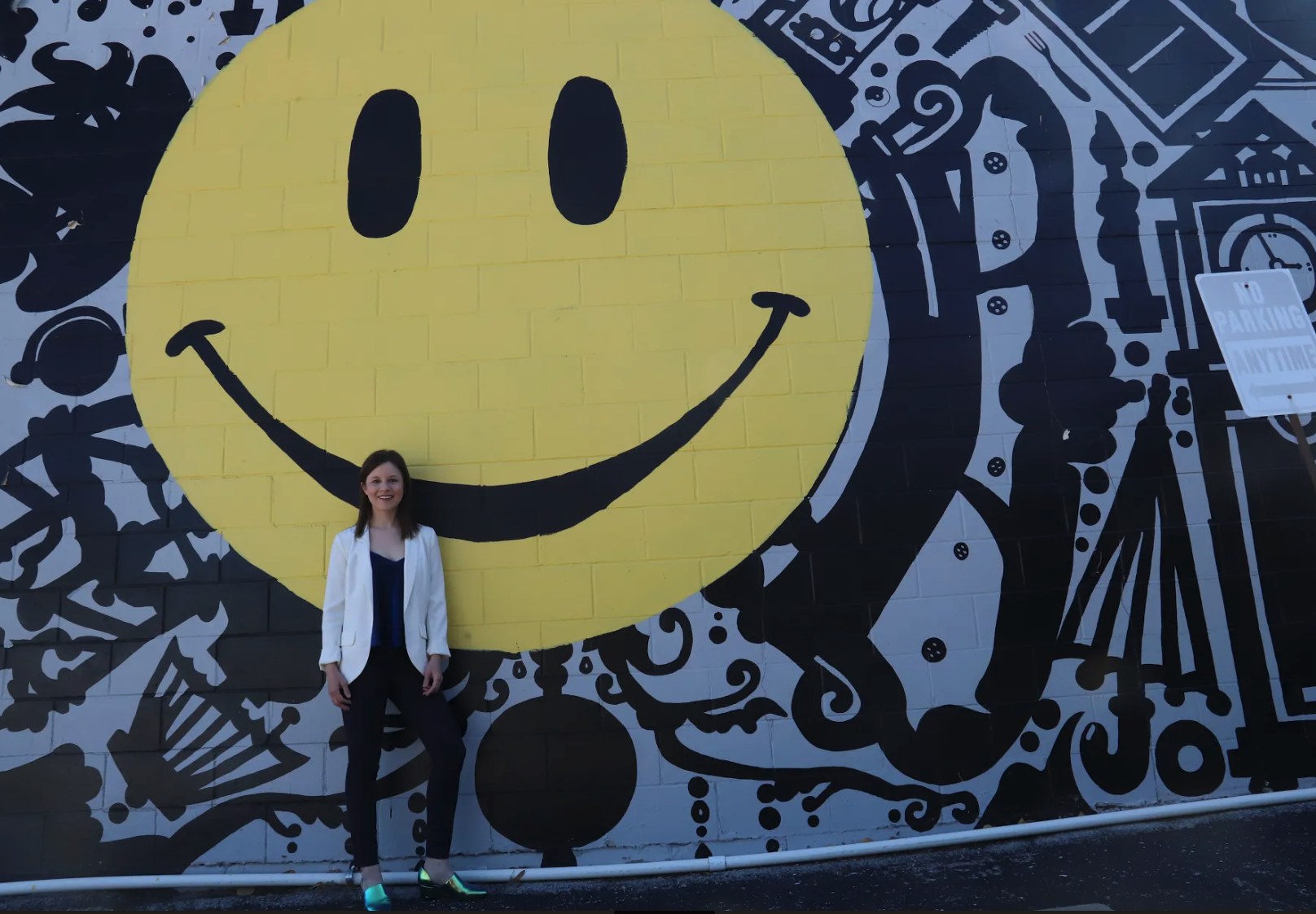A woman standing in front of a large mural of a yellow smiley face with black eyes and a big smile on a black and white background. She is wearing a white blazer, black pants, and teal shoes, and is smiling.
