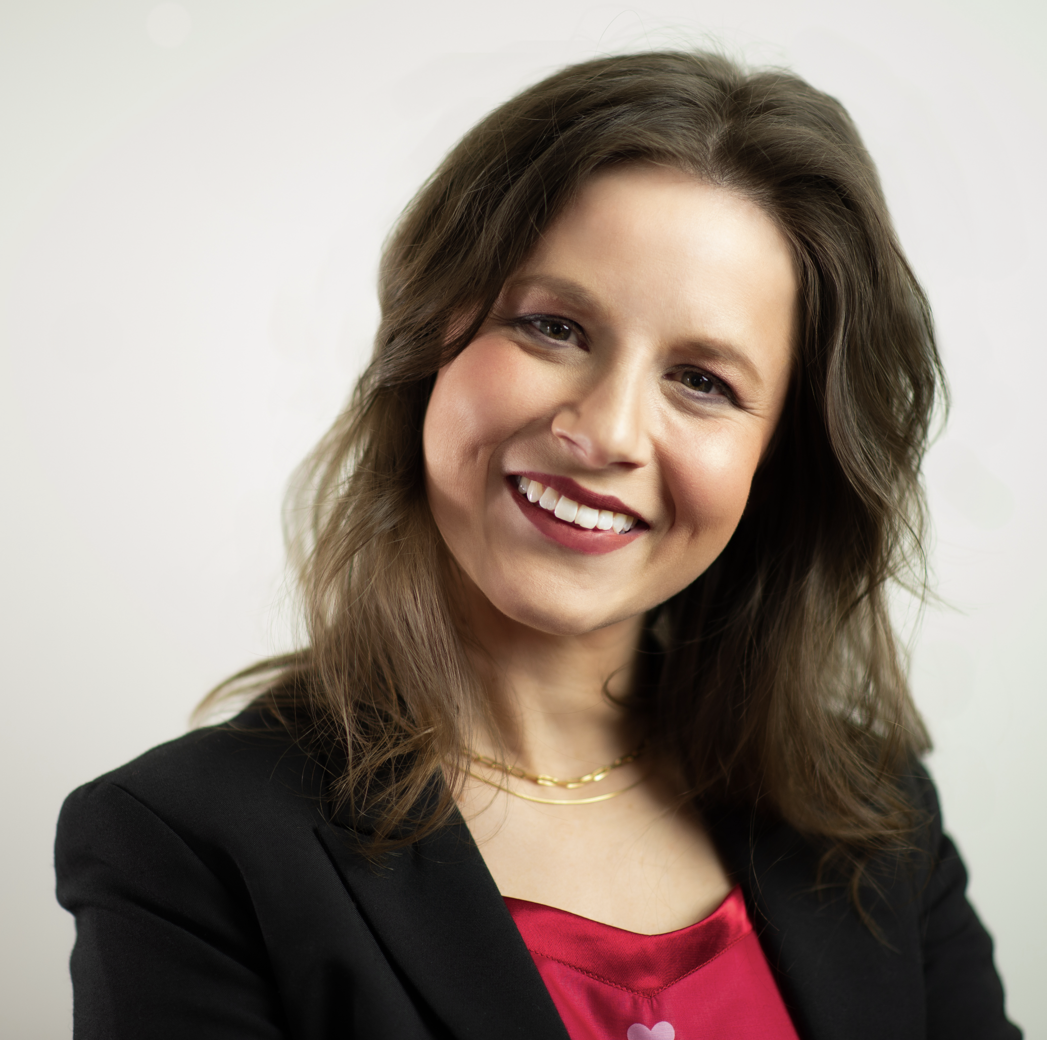 Close-up of a woman's face, showing her smiling with white teeth, fair skin, brown hair, wearing a black blazer, pink top with white hearts, and layered gold necklaces.
