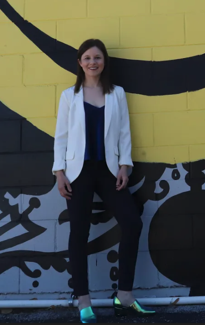 Woman standing in front of a colorful mural wall, wearing a white blazer, dark pants, and metallic shoes, smiling at the camera.