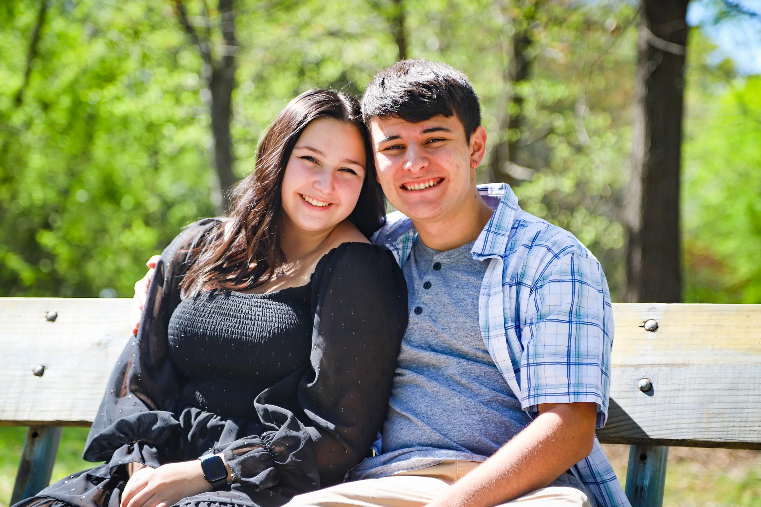 A young couple sitting on a park bench smiling, with trees and green foliage in the background on a sunny day.