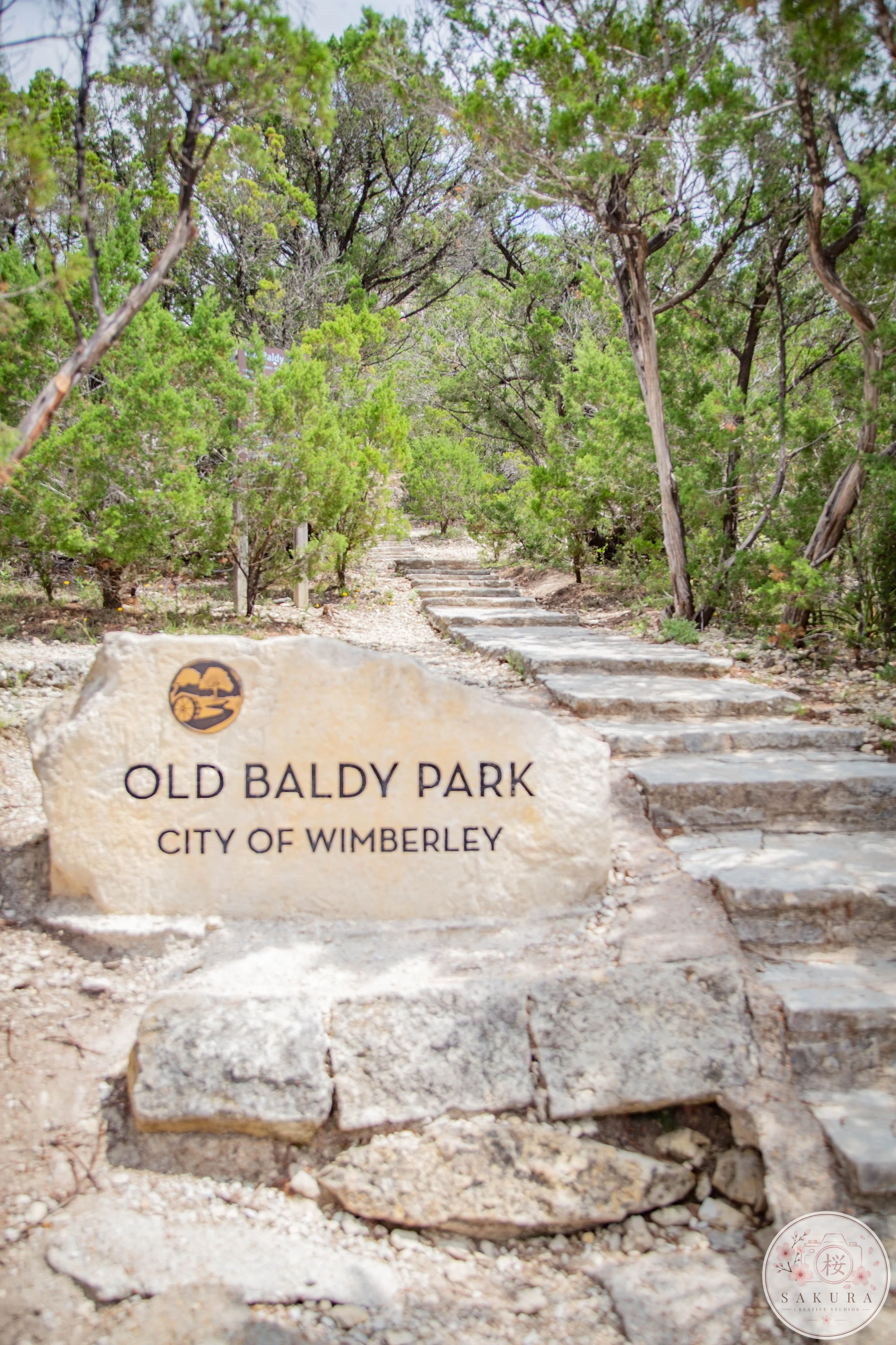 A stone marker in Old Baldy Park, City of Wimberley, with a park logo, surrounded by a dirt pathway and green trees with dense foliage