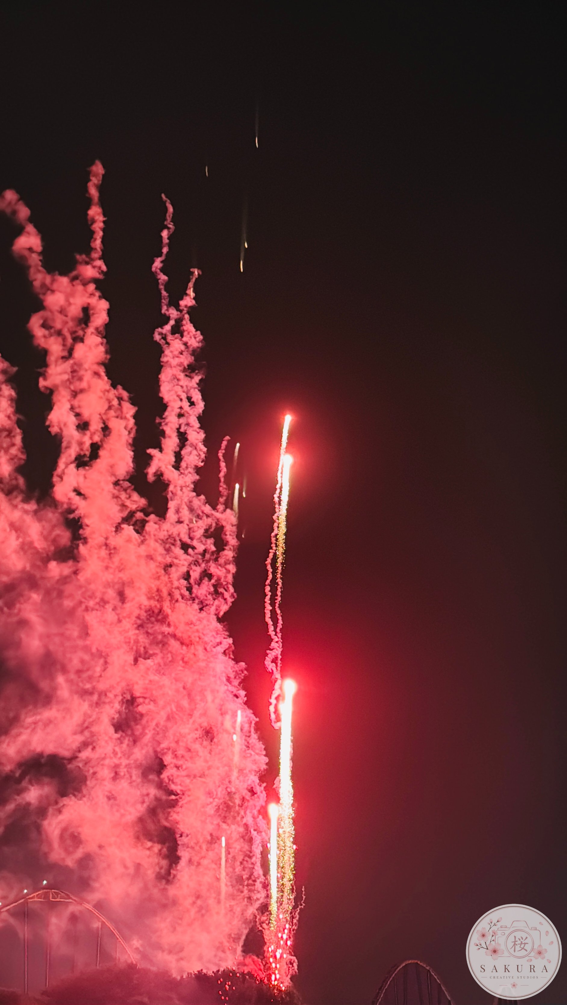 Fireworks with red smoke and streaks of light across the night sky, with some silhouettes of amusement park rides visible at the bottom.