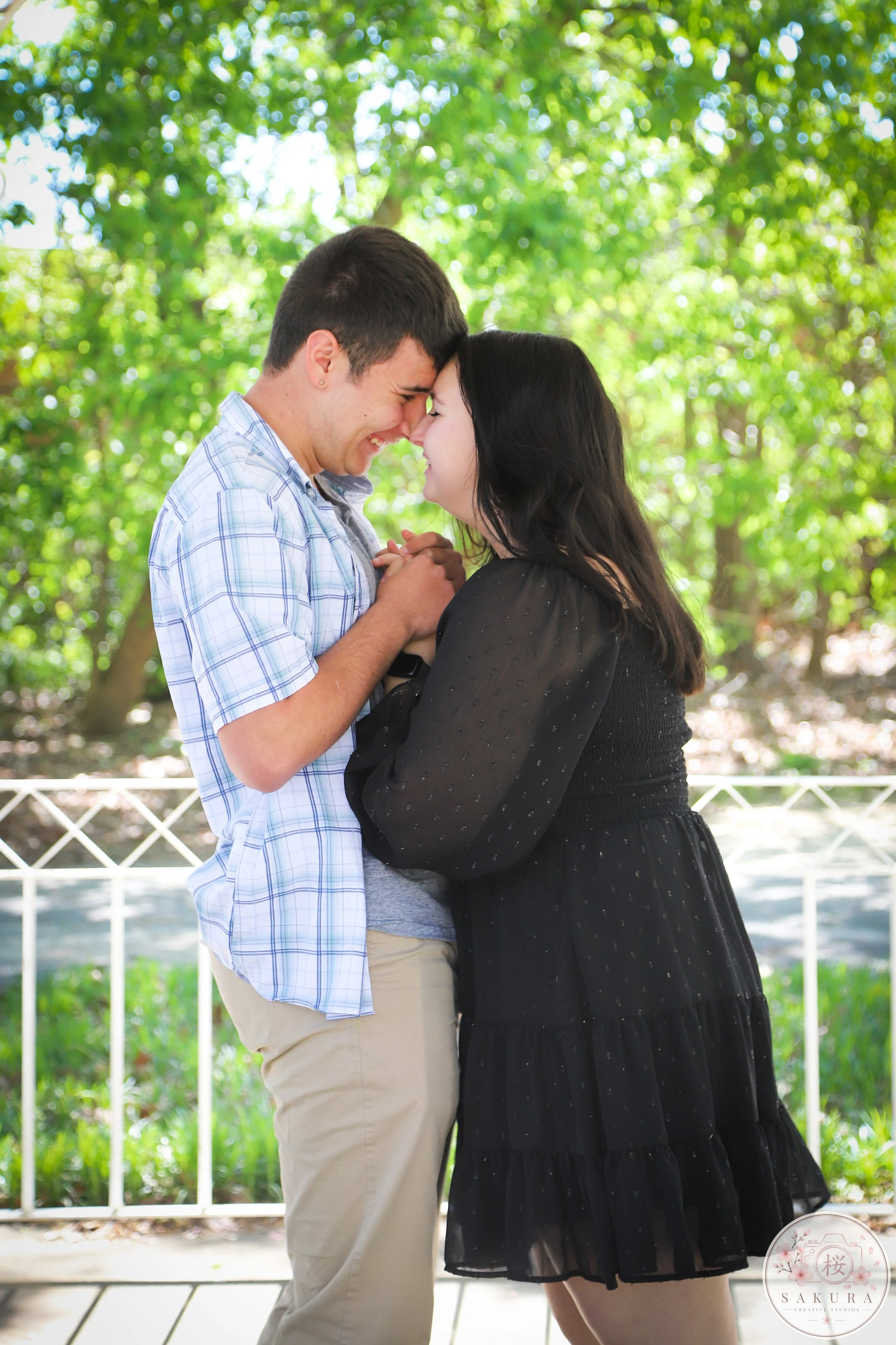A young couple standing outdoors, understanding each other, with their foreheads touching and smiling, surrounded by greenery.