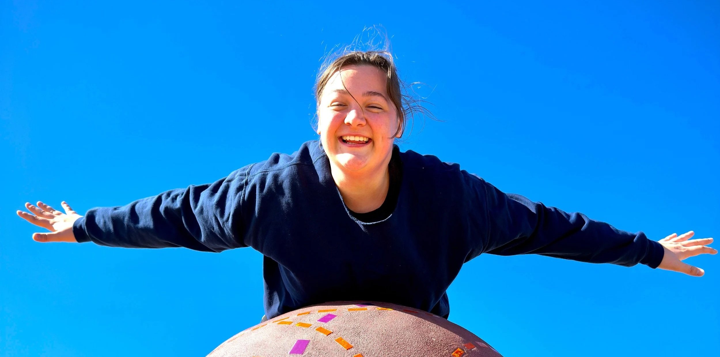 A young woman with brown hair smiling and extending her arms outward, standing outdoors against a clear blue sky.