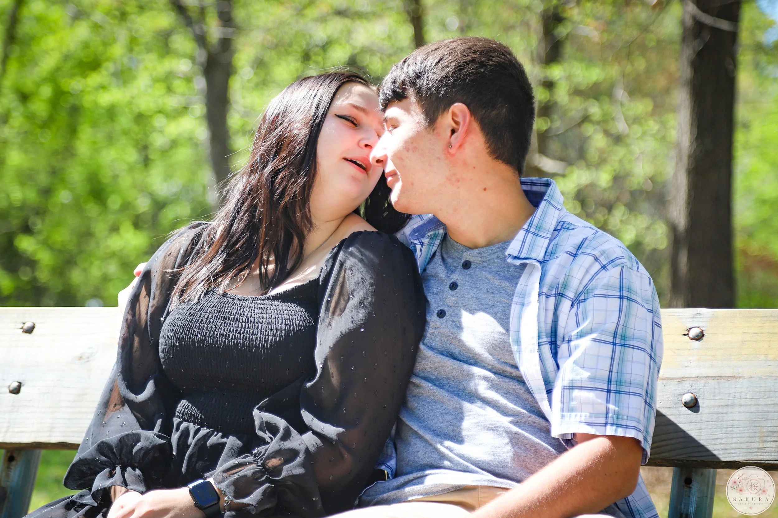 A young couple sitting on a wooden park bench, leaning in close and touching foreheads in an affectionate moment. They are outdoors with lush green trees in the background on a sunny day.