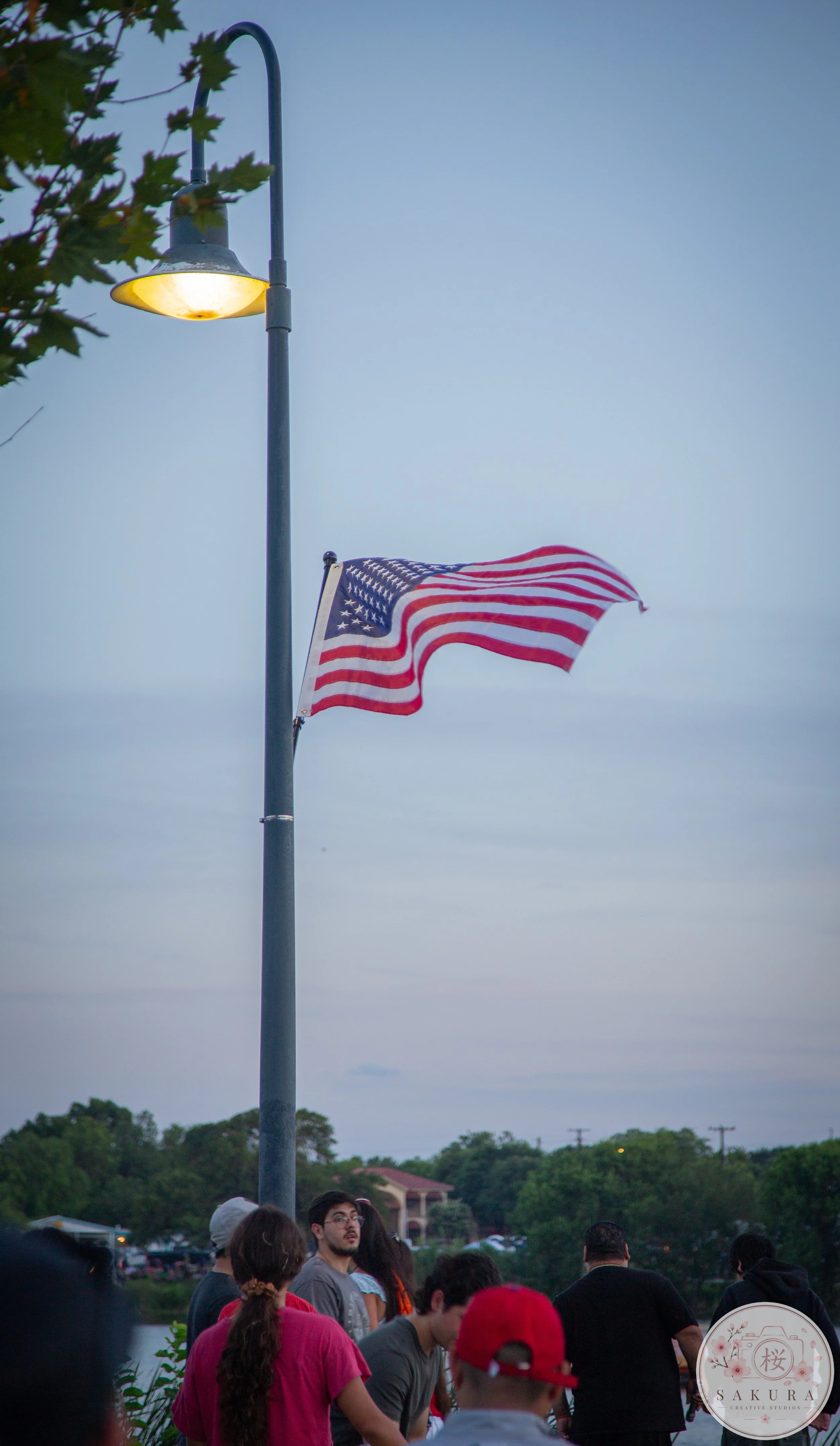 A Japanese flag waving near a street lamp with people gathered outdoors and trees in the background during dusk.