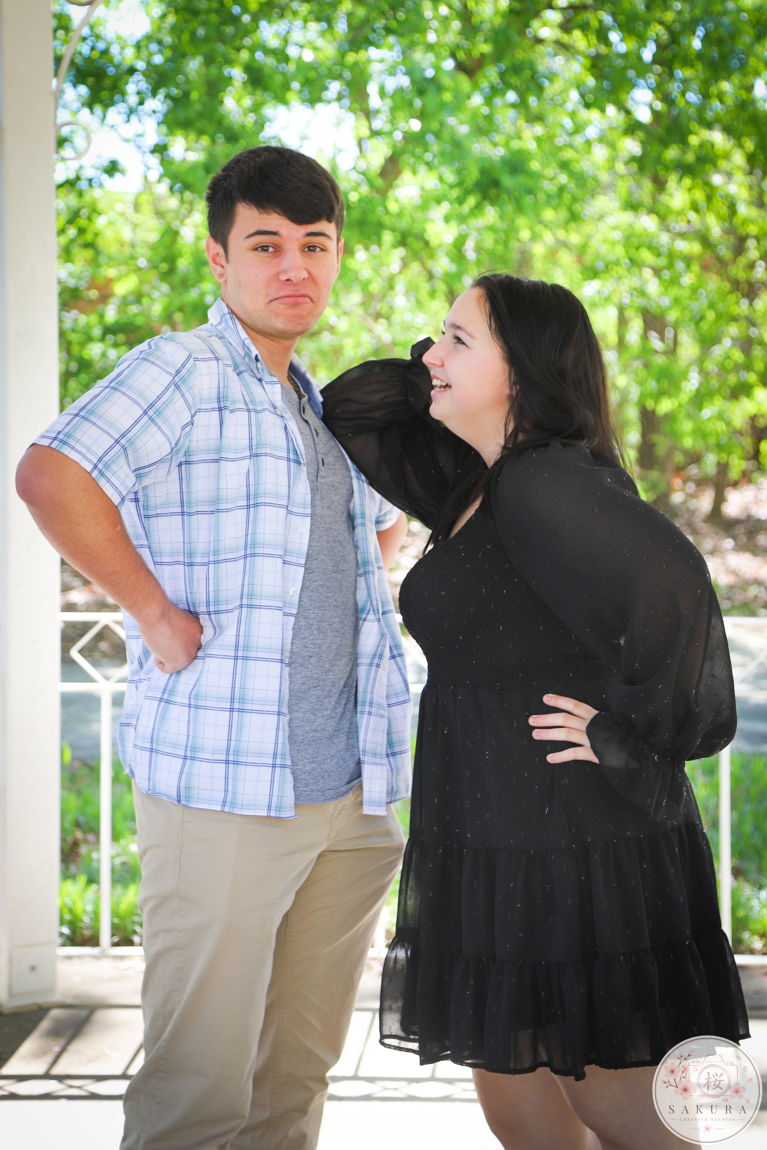 A young man with dark hair and a young woman with dark hair standing outdoors on a porch, with greenery and trees in the background. The woman is smiling and looking up at the man, who has a neutral expression. The man is wearing a light blue plaid s