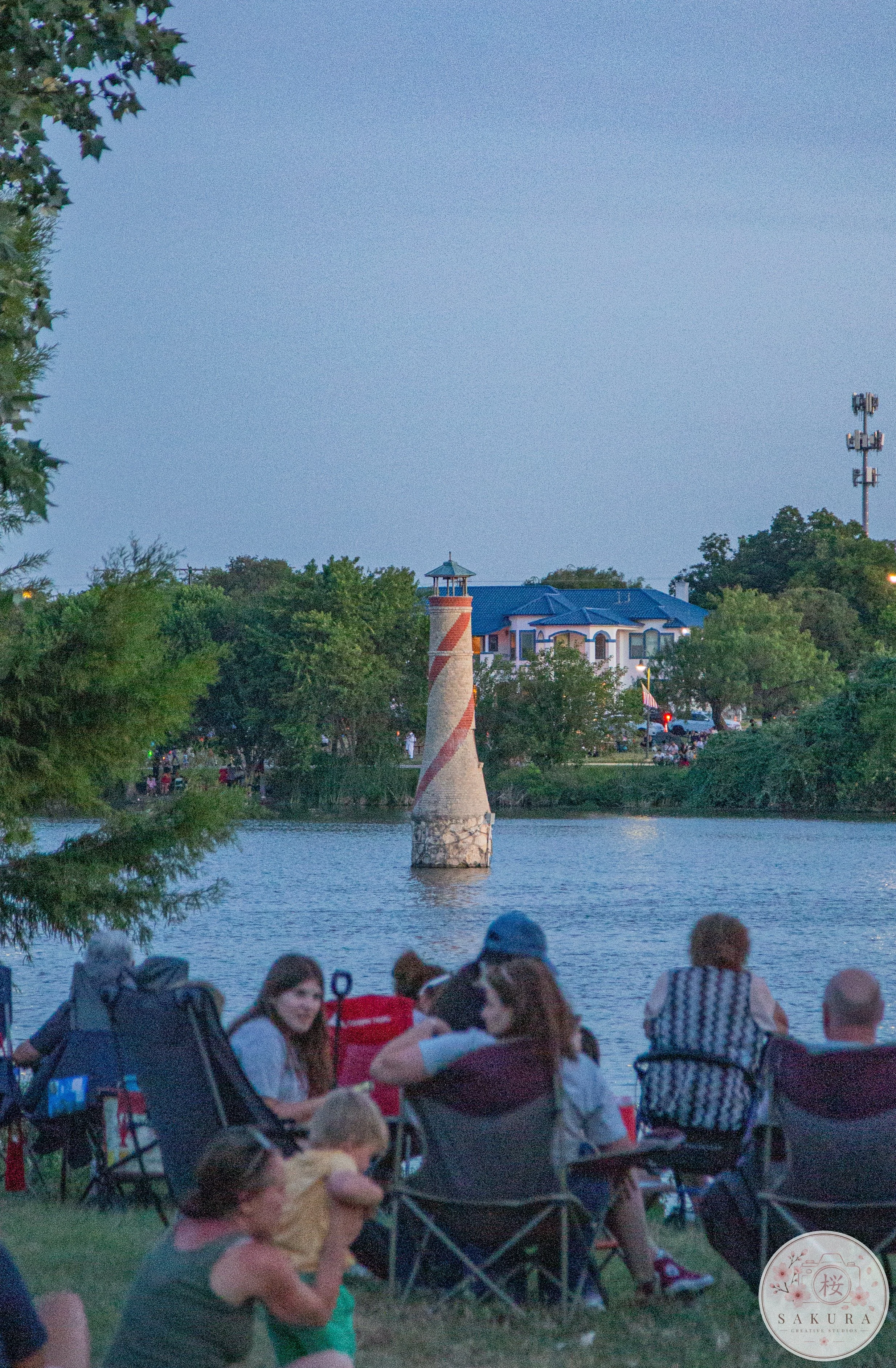 People sitting on the grass near a river, facing a lighthouse and a house with a blue roof in the distance, with trees and a communication tower in the background during dusk.