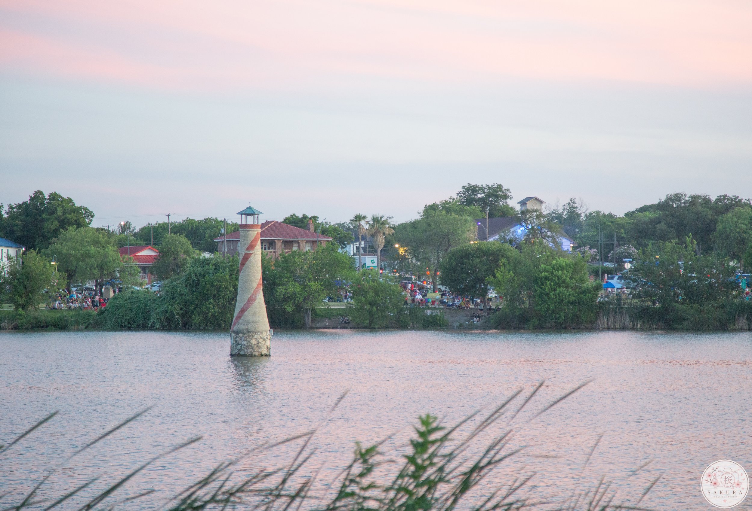 A body of water with a chimney-like structure in the middle. On the opposite side, there are buildings, trees, and a crowd of people, possibly at a park or festival during dusk.