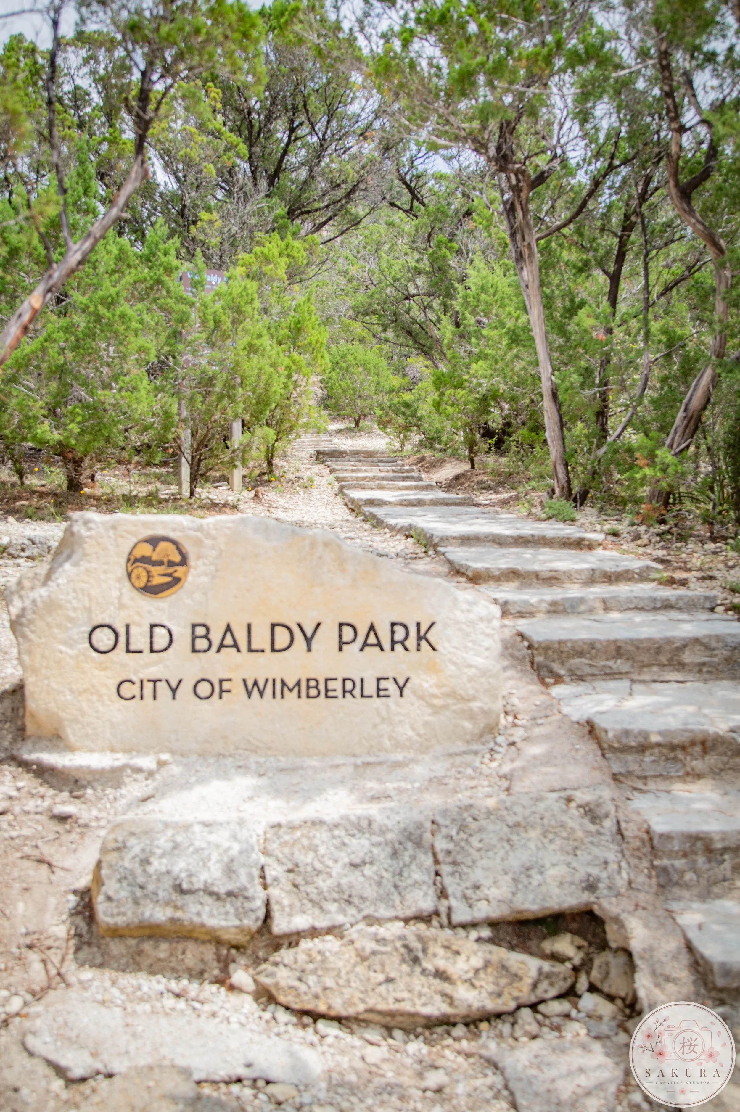 A stone sign with the inscription "Old Baldy Park, City of Wimberley" in front of a dirt pathway with stone steps surrounded by green trees.