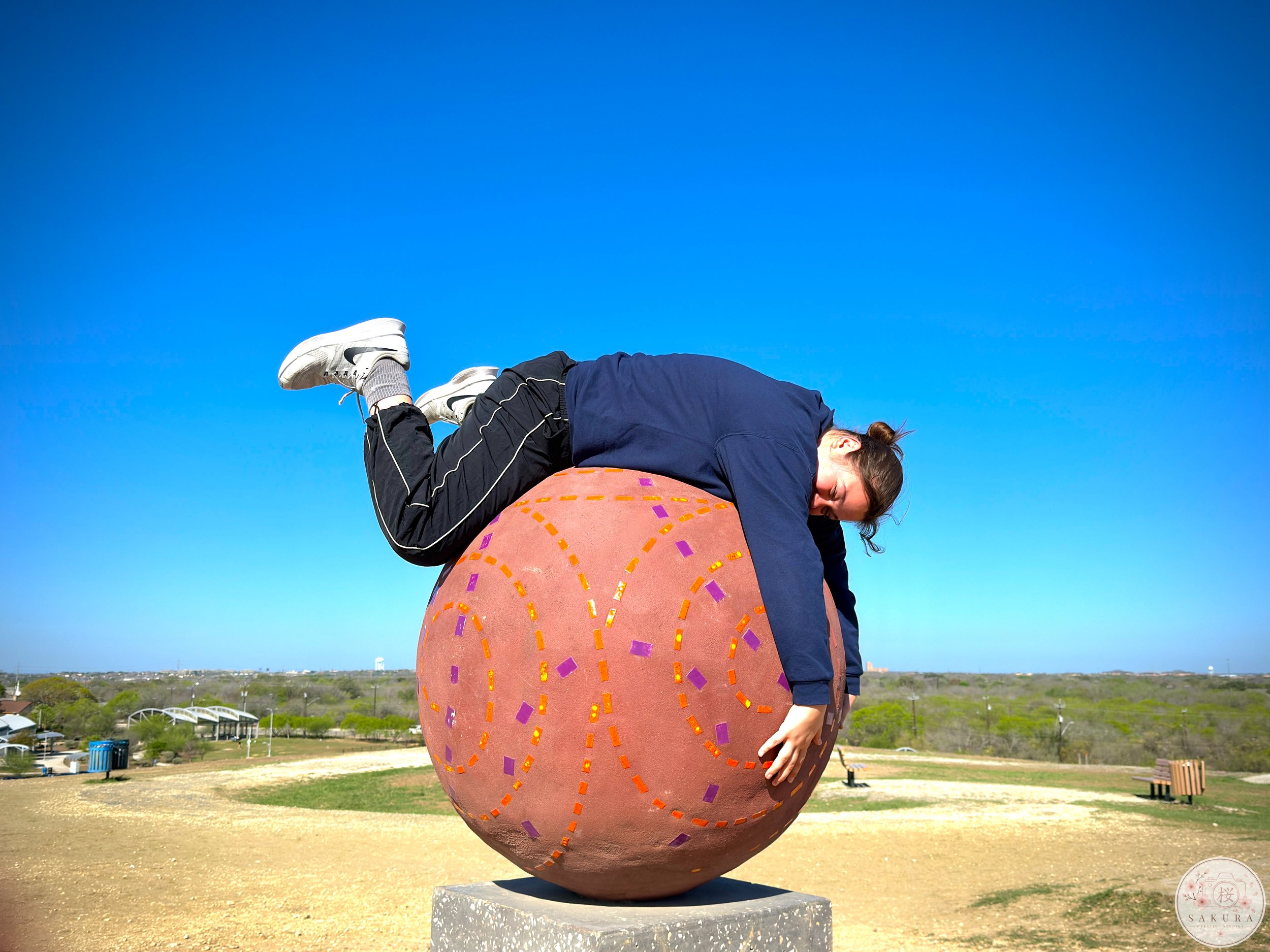 Person with brown hair wearing a navy jacket and gray sneakers, balancing on a large, colorful sculpture resembling a decorated fruit or vegetable, outdoors under a clear blue sky.