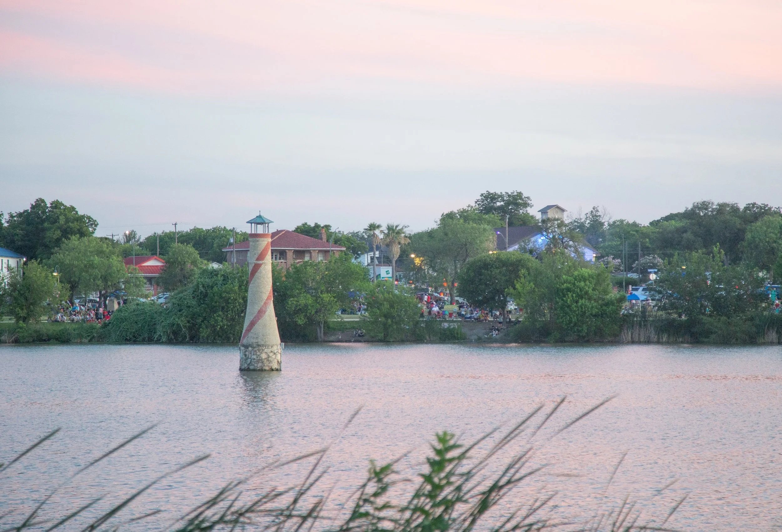 View of a body of water with grass in the foreground, a lighthouse-like structure in the water, houses and trees on the opposite shore, and a partly cloudy sky at dusk.